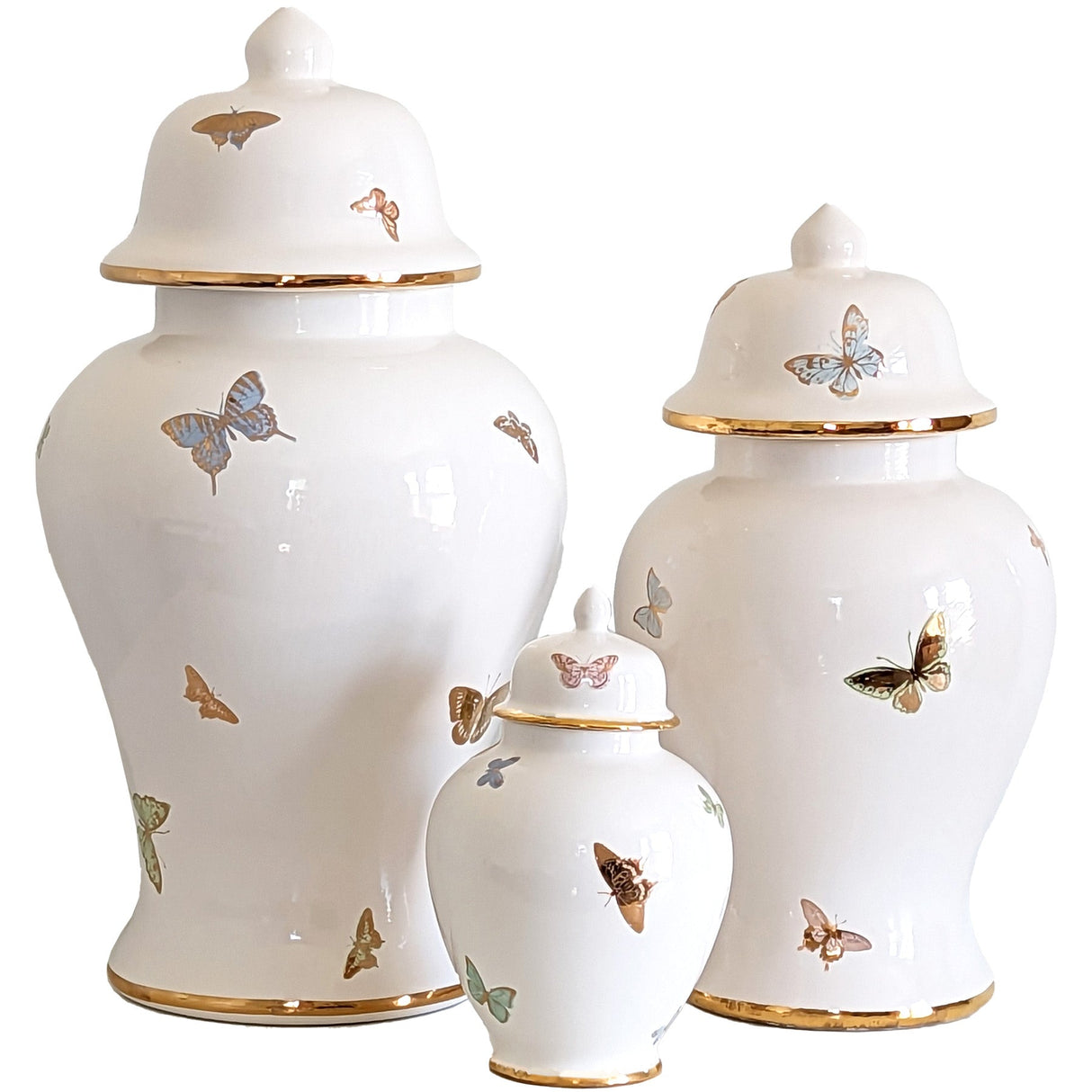 A set of three white ginger jars with gold trim and multicolored butterflies painted on them with more fold details, in varying sizes.