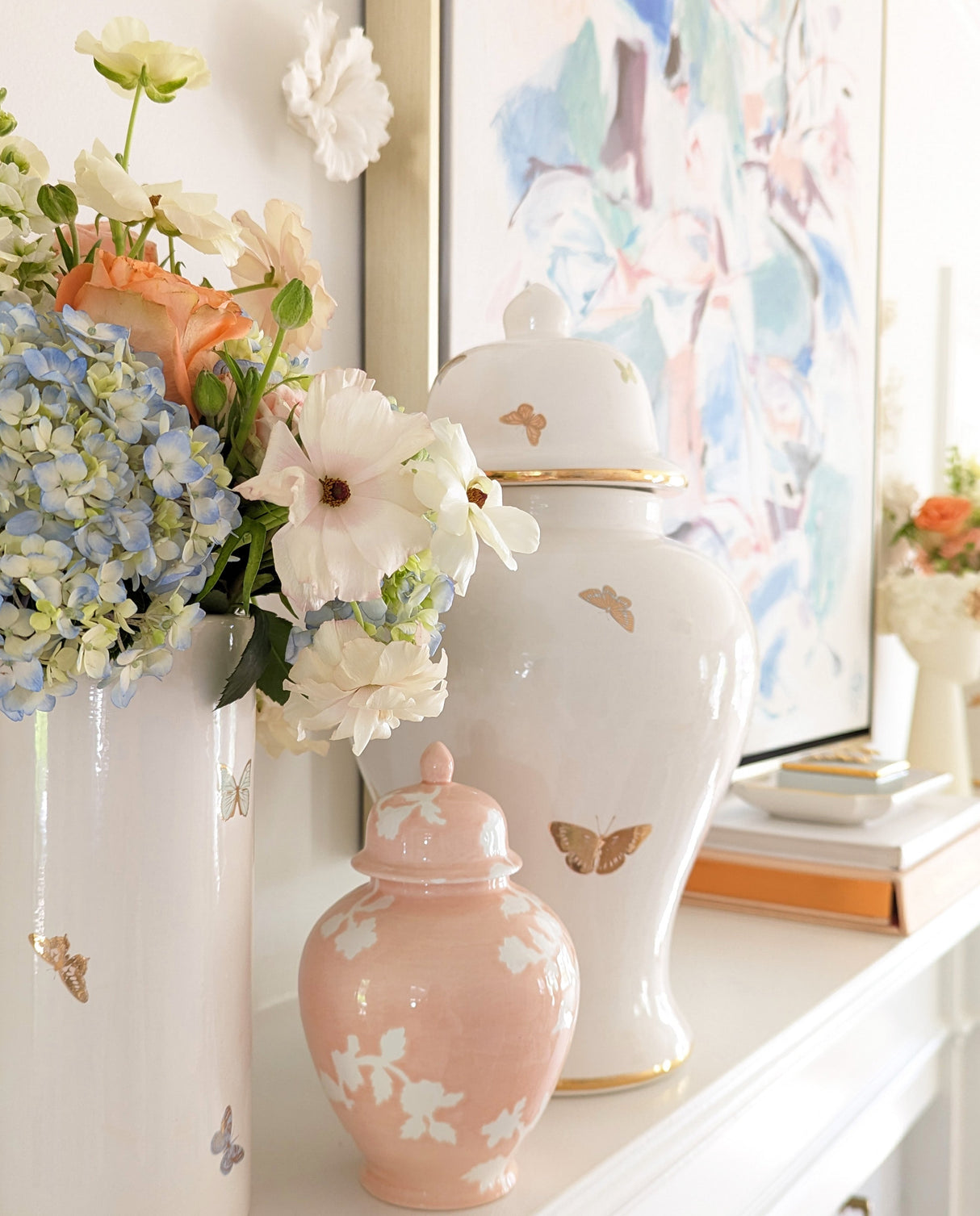 Pastel flower arrangement in a white butterfly-decorated vase beside blush and white ceramic ginger jars on a white mantel with an abstract pastel painting in the background.
