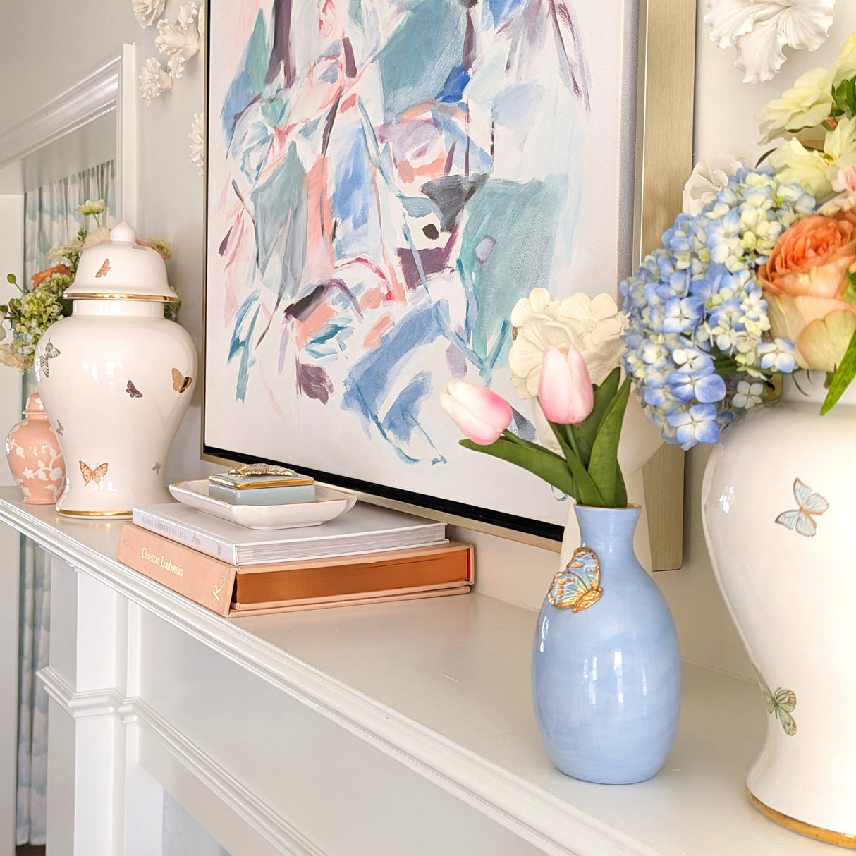 A white mantel styled with a pastel abstract painting, butterfly-decorated ceramic vases, fresh flowers, stacked books, and a blue vase holding pink tulips.
