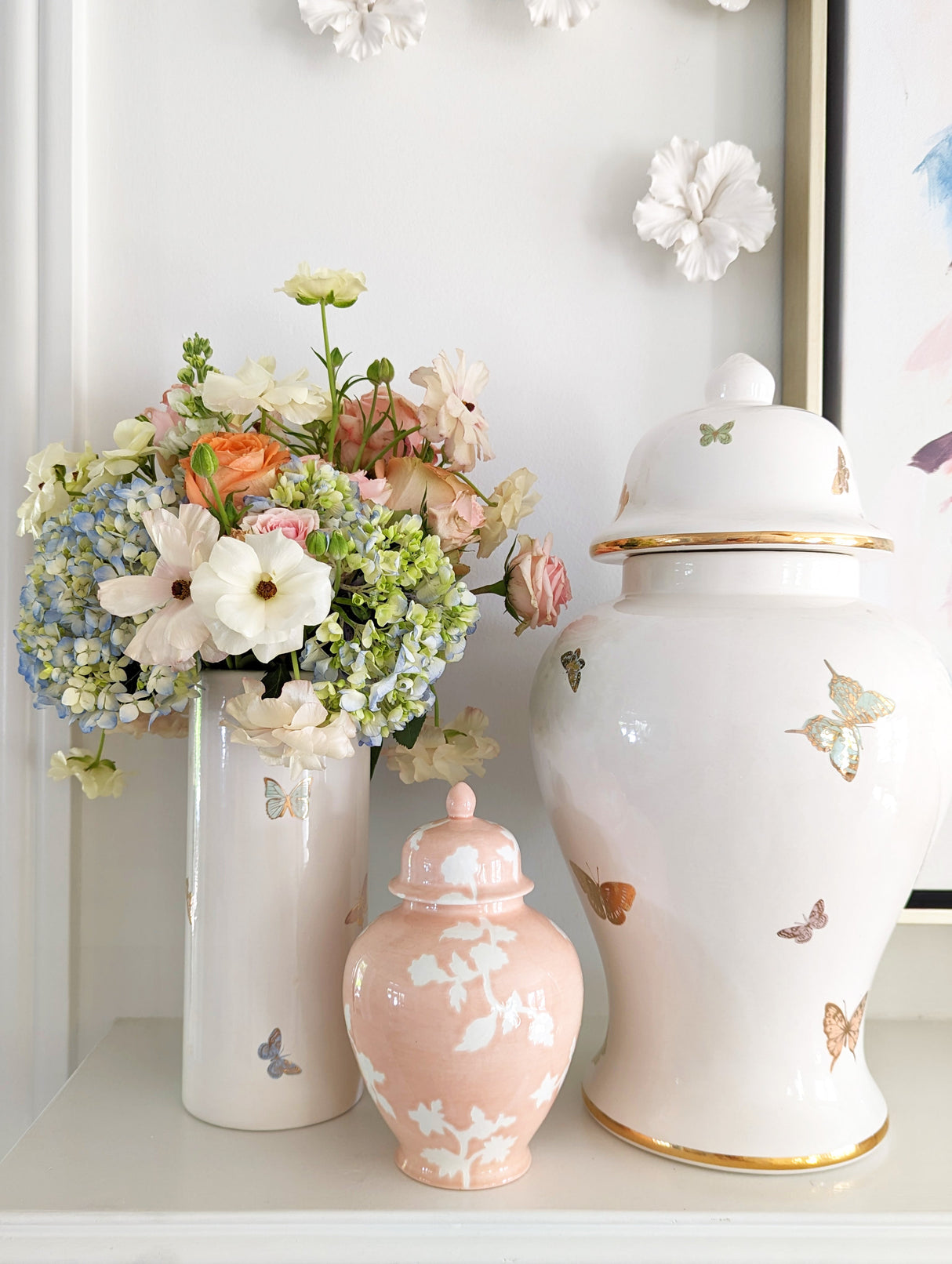 Pastel flower arrangement in a white multi colored and golden butterfly-decorated vase beside blush and white ceramic ginger jar decorated with matching butterflies on a white shelf.